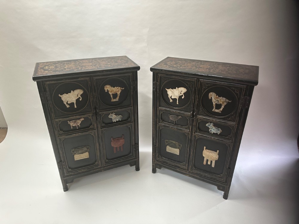 A pair of black lacquered two-door Chinoiserie cabinets featuring carved hardstone inlays of horses, animals, and vessels, topped with gilt scrollwork motifs. View V