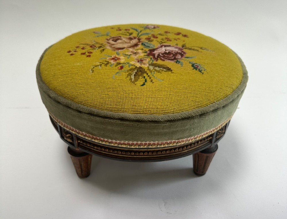 A low circular French footstool featuring a floral needlepoint seat in mustard yellow with green velvet trim and carved wooden legs.