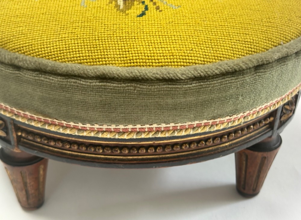 A low circular French footstool featuring a floral needlepoint seat in mustard yellow with green velvet trim and carved wooden legs. View VI