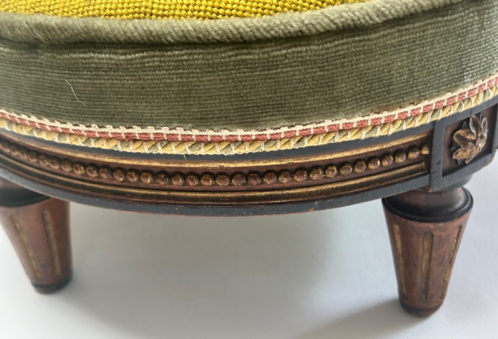 A low circular French footstool featuring a floral needlepoint seat in mustard yellow with green velvet trim and carved wooden legs. View VII