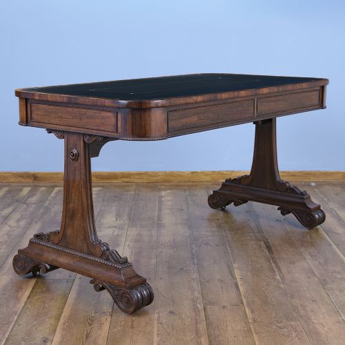 A Regency rosewood writing desk featuring a black leather inset surface, frieze drawers, and carved trestle end supports on brass castors. View X