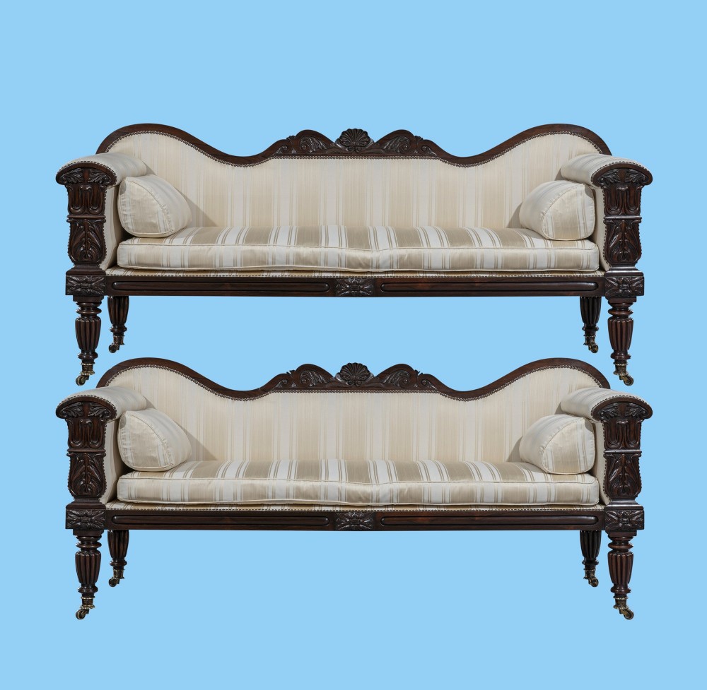 A matching pair of 19th-century dark rosewood sofas with carved cresting, scroll arms, reeded legs on brass castors, and cream striped fabric upholstery.