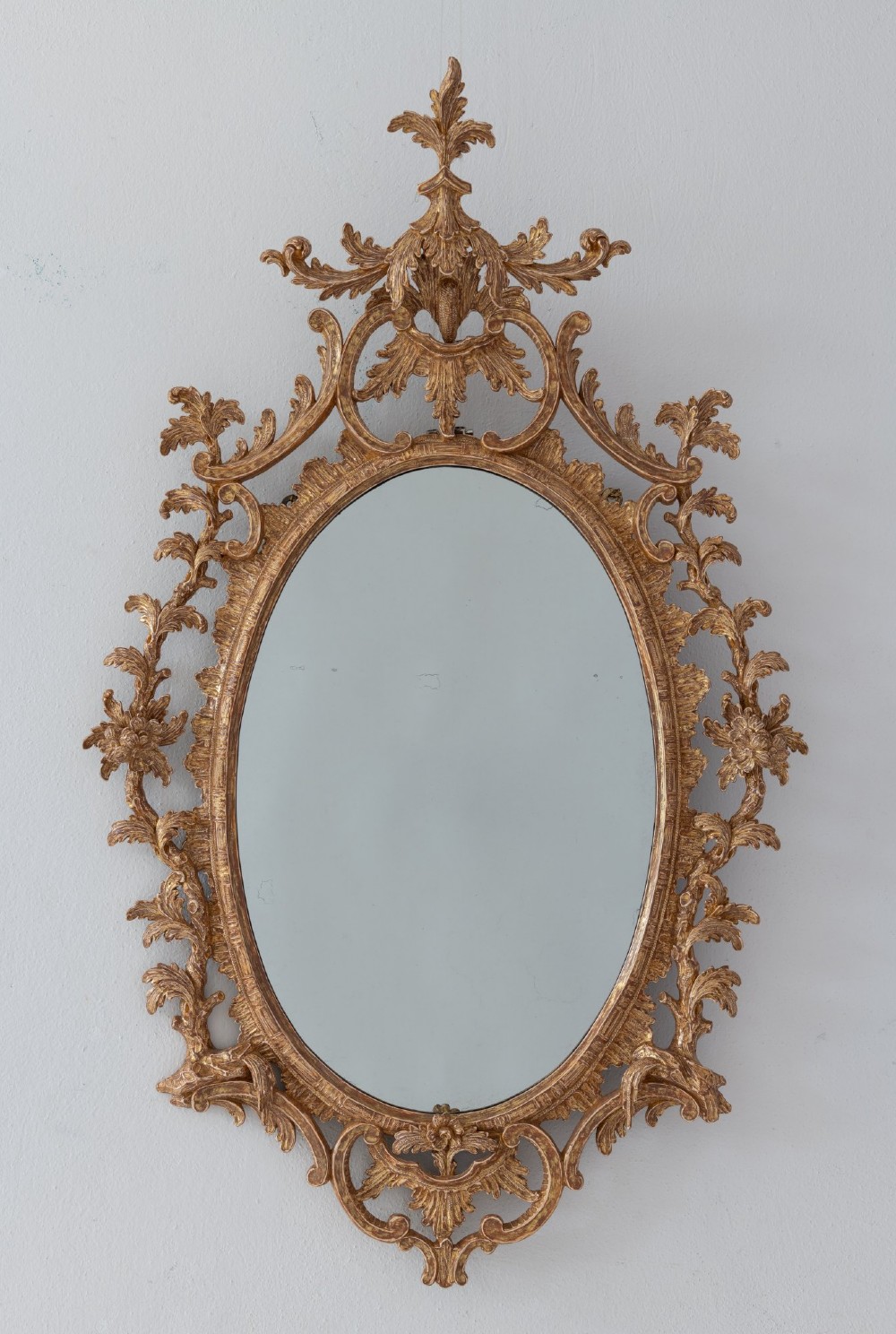 An 18th-century style oval wall mirror featuring a carved giltwood frame with detailed acanthus leaves, C-scrolls, and floral motifs.