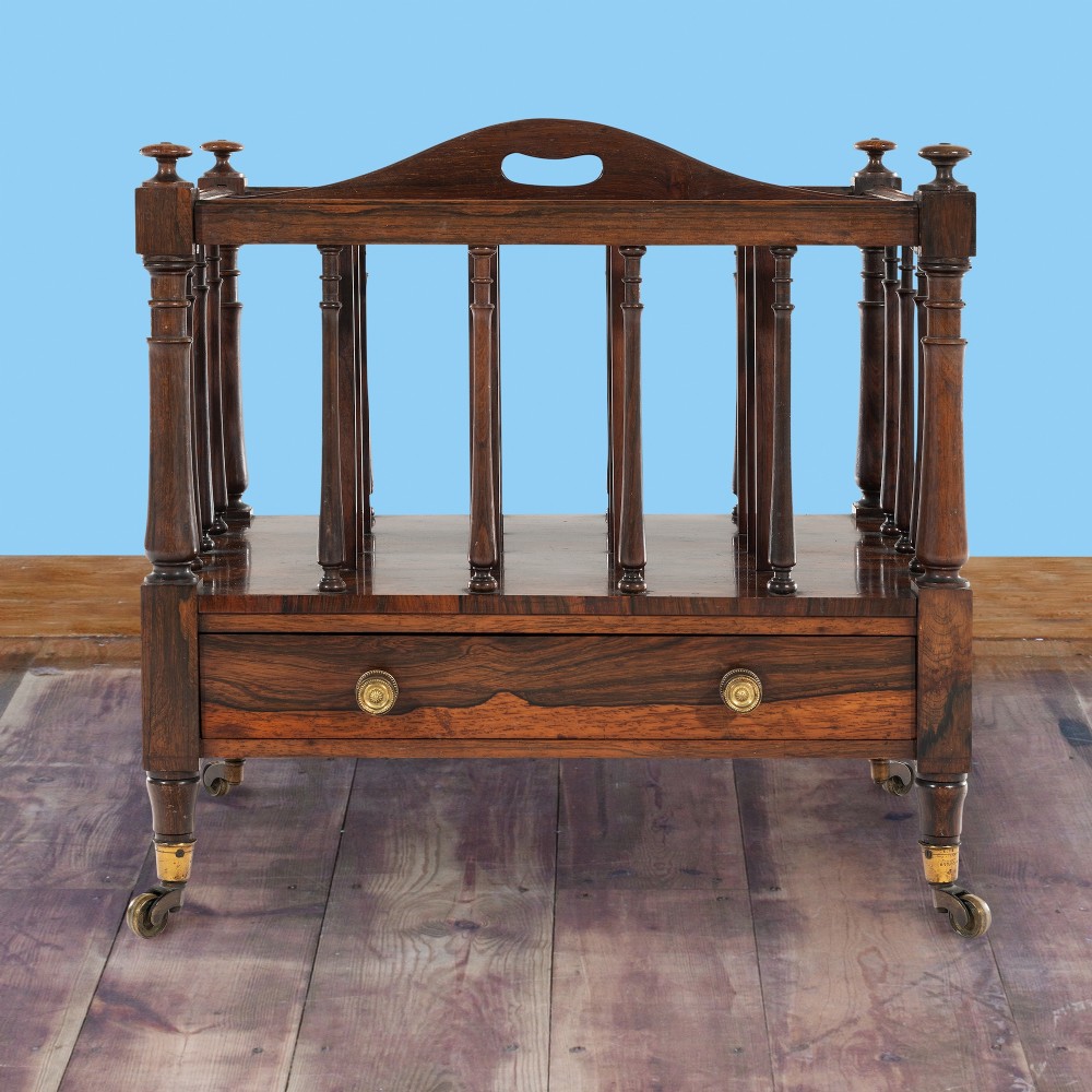 A Victorian rosewood music canterbury featuring turned spindle dividers, a top carrying handle, a lower drawer with two brass knobs, and turned legs with brass castors.