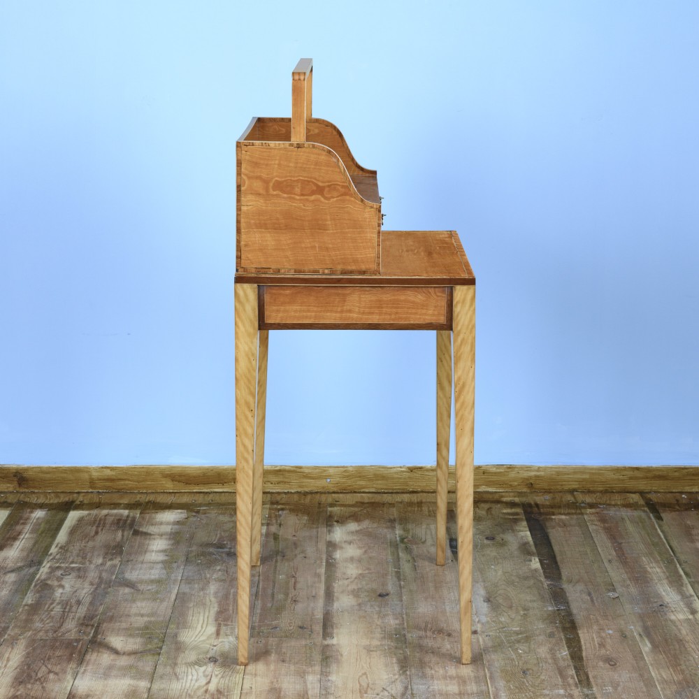 An 18th-century Georgian satinwood cheveret writing table featuring slender square tapered legs, brass ring pulls, and a upper cabinet with an integrated handle. View X