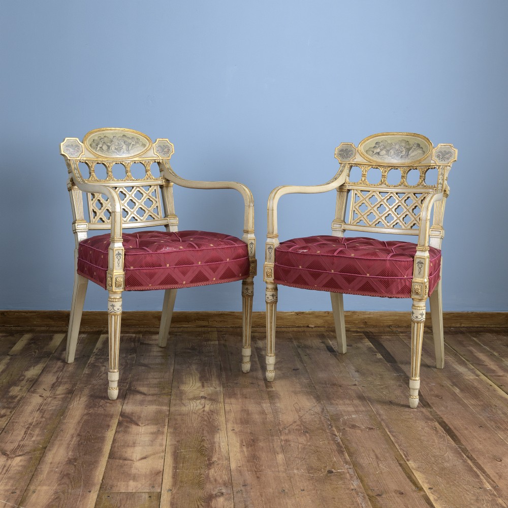 A pair of cream-painted and parcel-gilt Neoclassical armchairs featuring pierced lattice backs, painted oval medallions, and crimson fabric seats.