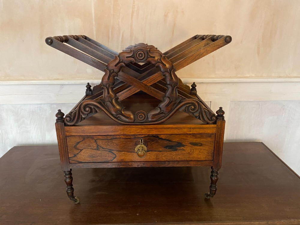 An antique dark rosewood Regency Canterbury featuring carved laurel scroll dividers, a base drawer with a brass ring handle, and turned legs with brass castors. View II