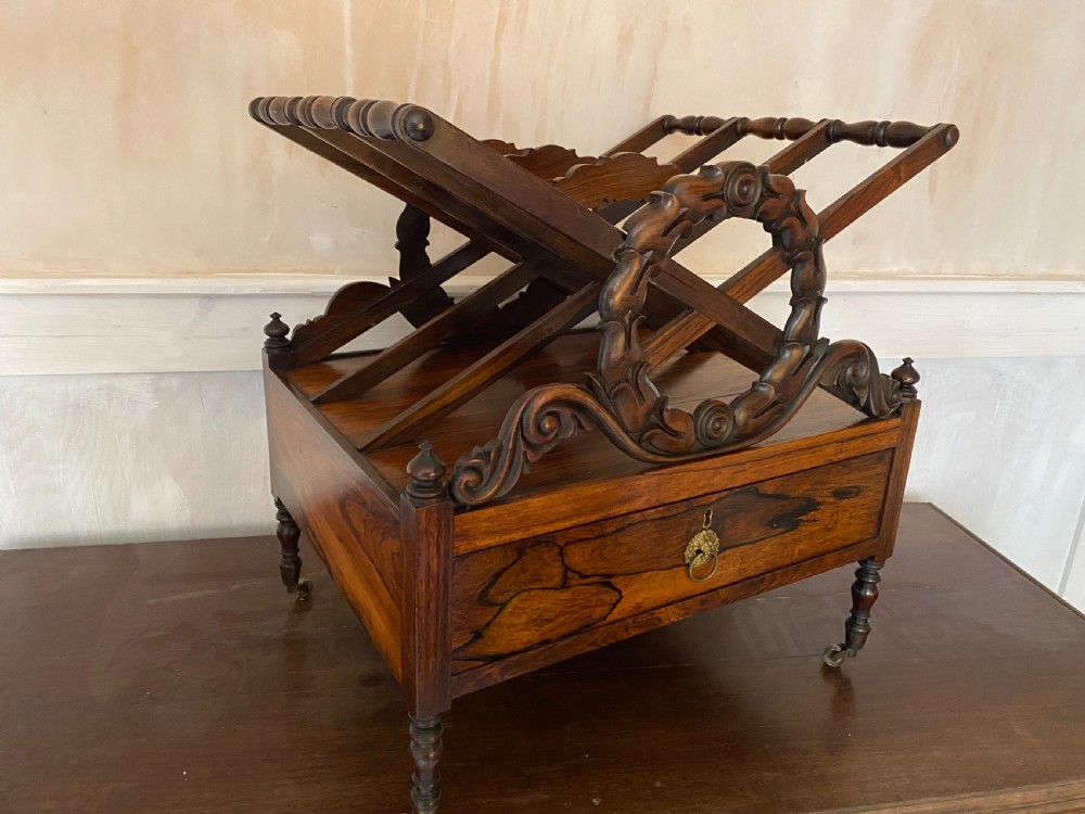 An antique dark rosewood Regency Canterbury featuring carved laurel scroll dividers, a base drawer with a brass ring handle, and turned legs with brass castors. View III