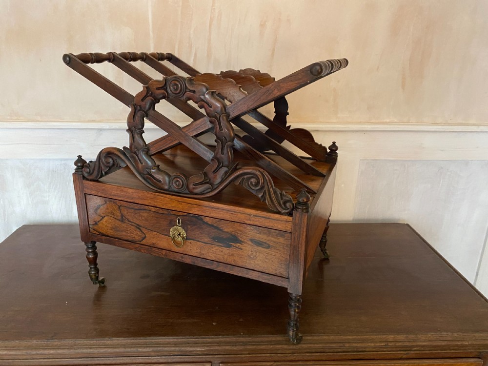 An antique dark rosewood Regency Canterbury featuring carved laurel scroll dividers, a base drawer with a brass ring handle, and turned legs with brass castors. View IV