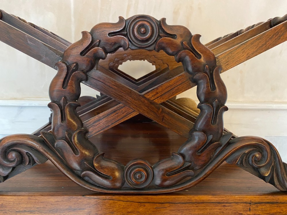 An antique dark rosewood Regency Canterbury featuring carved laurel scroll dividers, a base drawer with a brass ring handle, and turned legs with brass castors. View V