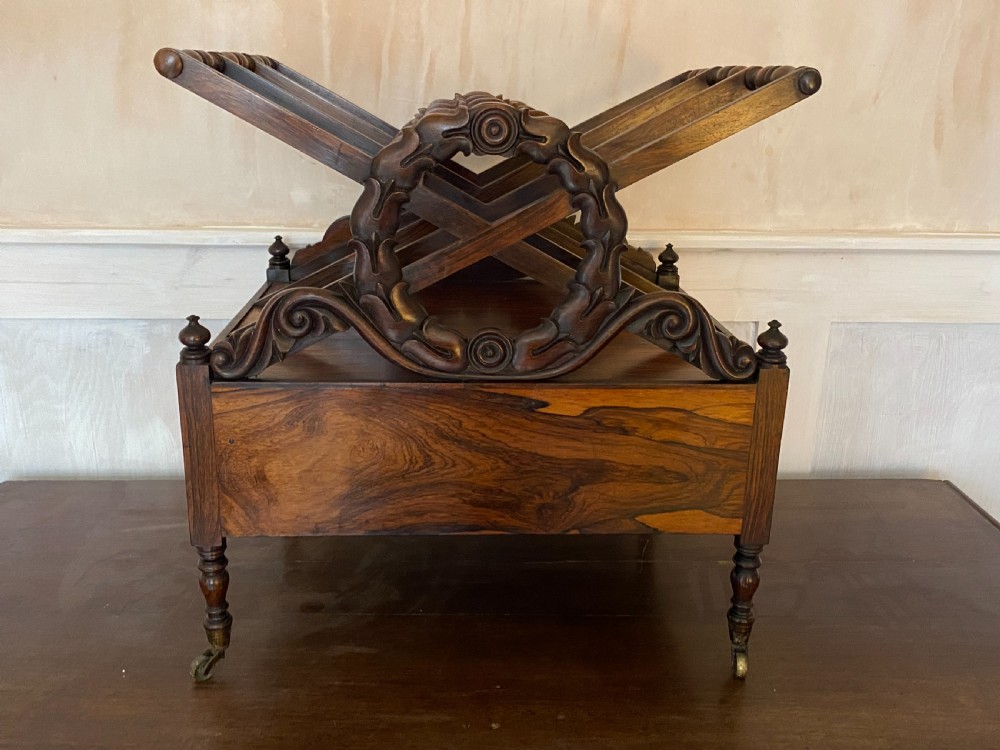 An antique dark rosewood Regency Canterbury featuring carved laurel scroll dividers, a base drawer with a brass ring handle, and turned legs with brass castors. View XIV