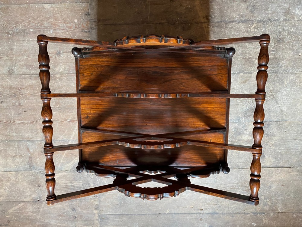 An antique dark rosewood Regency Canterbury featuring carved laurel scroll dividers, a base drawer with a brass ring handle, and turned legs with brass castors. View XXI