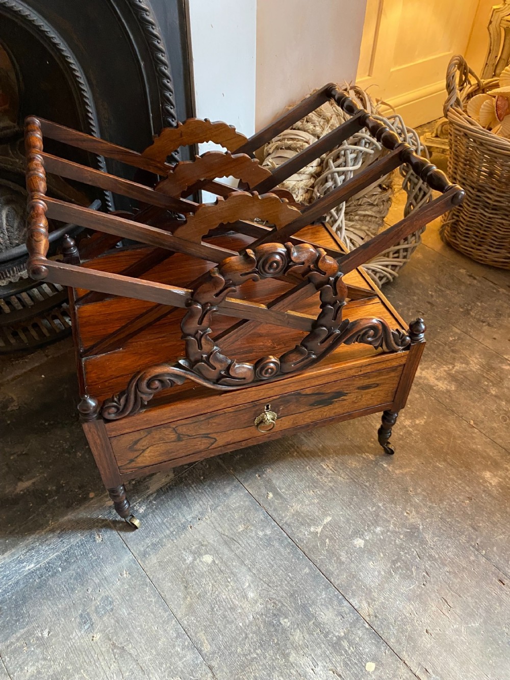 An antique dark rosewood Regency Canterbury featuring carved laurel scroll dividers, a base drawer with a brass ring handle, and turned legs with brass castors.