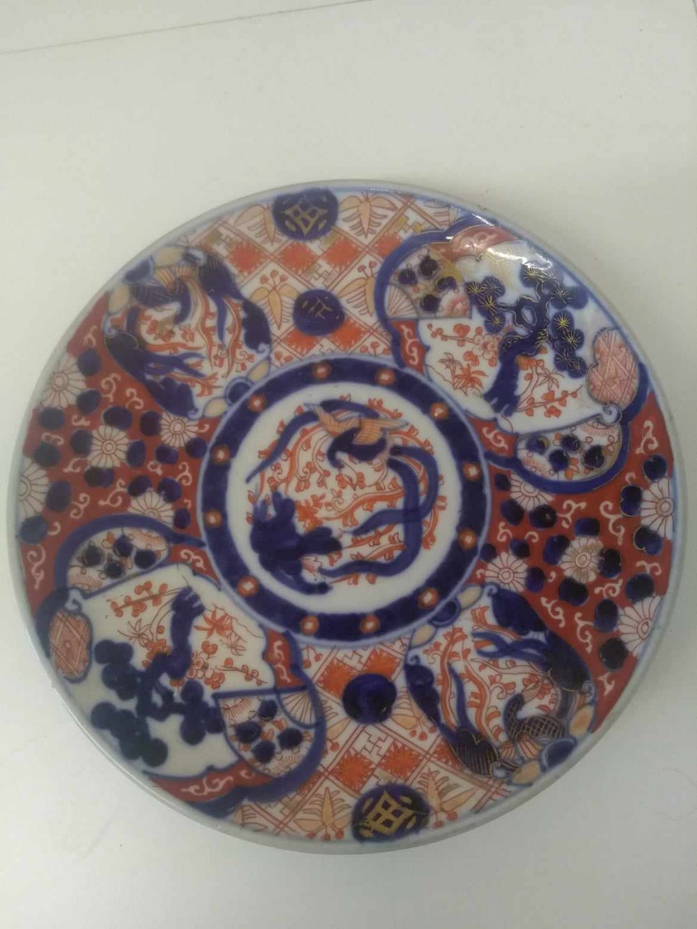A circular Japanese Imari porcelain plate decorated with hand-painted reserves containing floral motifs, pine trees, and a central phoenix medallion in cobalt blue, iron red, and gold accents.