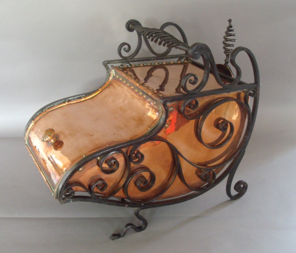 An Arts and Crafts copper coal purdonium resting within an ornamental black wrought iron scrollwork cradle with a twisted handle.