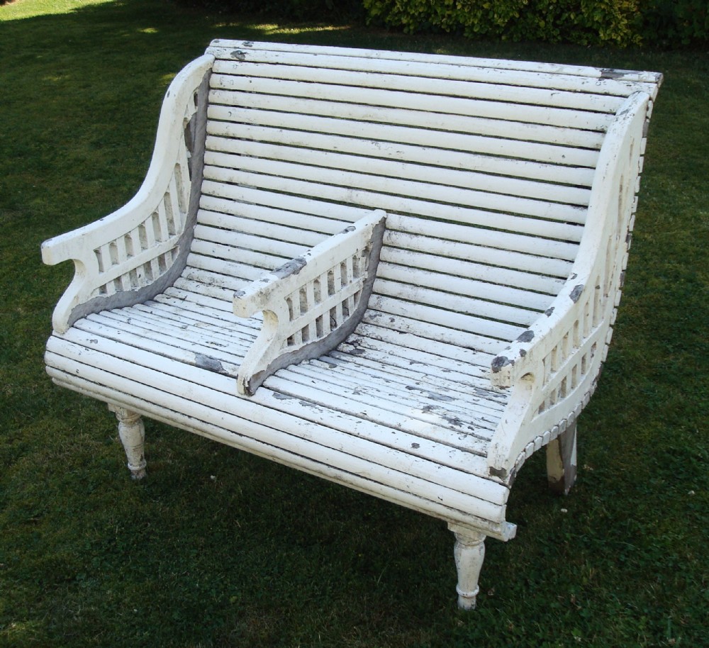 A distressed white painted slatted teak garden seat featuring a curved backrest, central divider armrest, and lattice detailed sides.