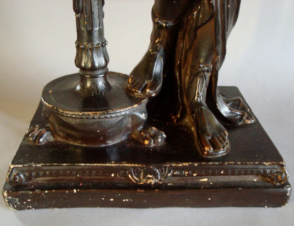 A dark bronzed plaster sculpture depicting a draped classical female figure standing beside a decorative column on a rectangular pedestal. View VII