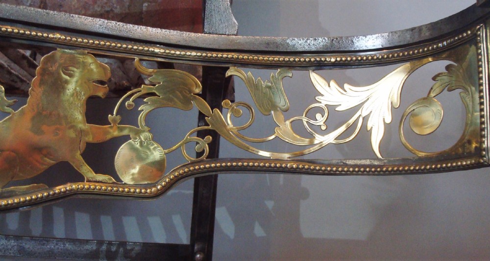 A 19th-century Neoclassical style fire grate constructed from black iron with a scroll-ended basket, flanked by polished brass columns and an ornate pierced brass apron featuring lion motifs. View X