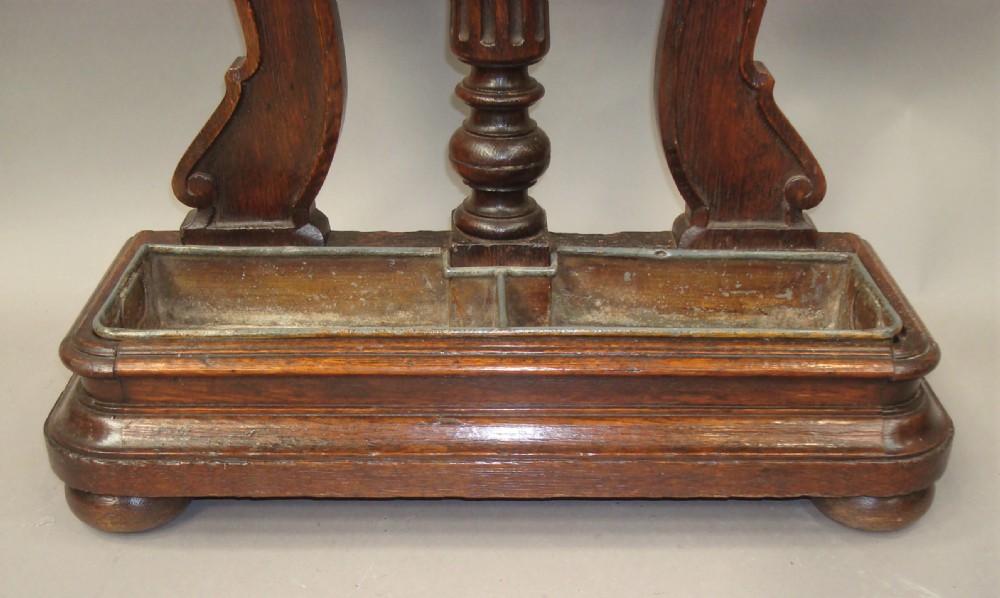An antique dark oak stick stand featuring scroll support arms, a carved top cresting, a central column, and a removable metal drip tray at the base. View IX