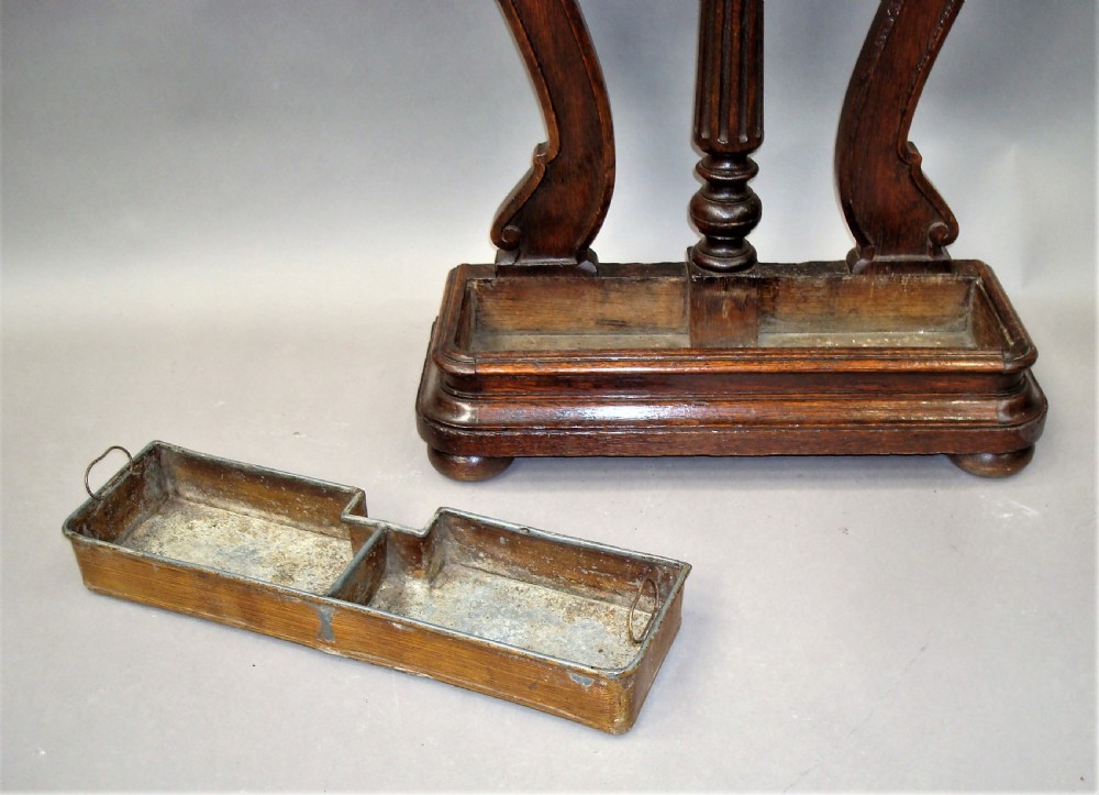 An antique dark oak stick stand featuring scroll support arms, a carved top cresting, a central column, and a removable metal drip tray at the base. View X