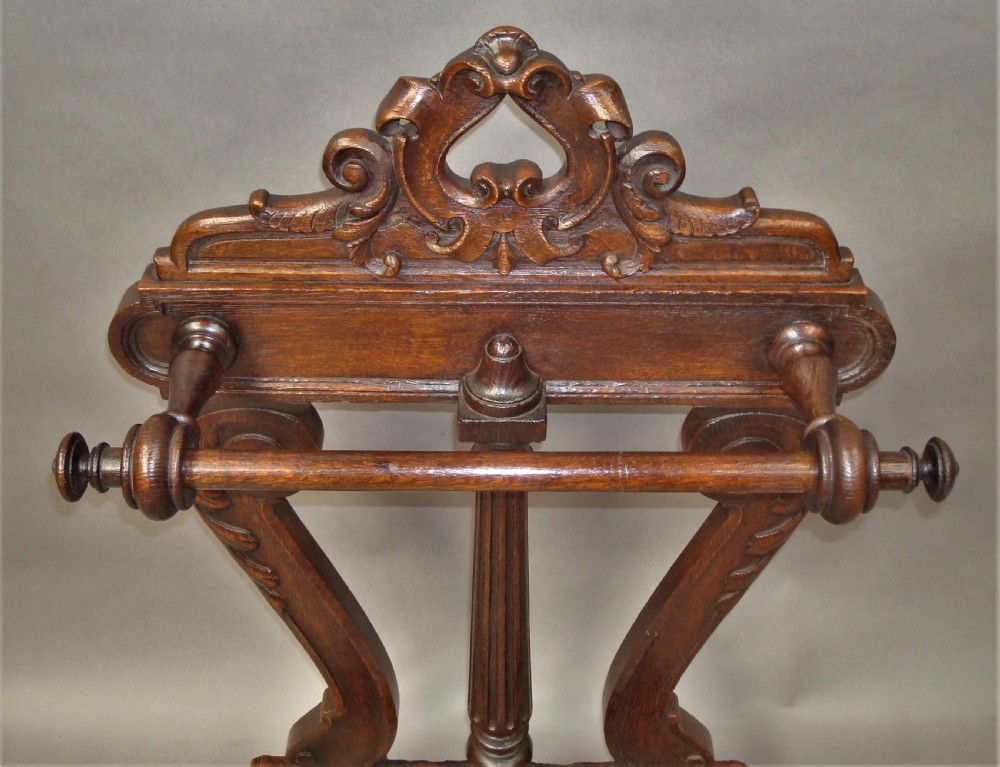 An antique dark oak stick stand featuring scroll support arms, a carved top cresting, a central column, and a removable metal drip tray at the base. View XIII