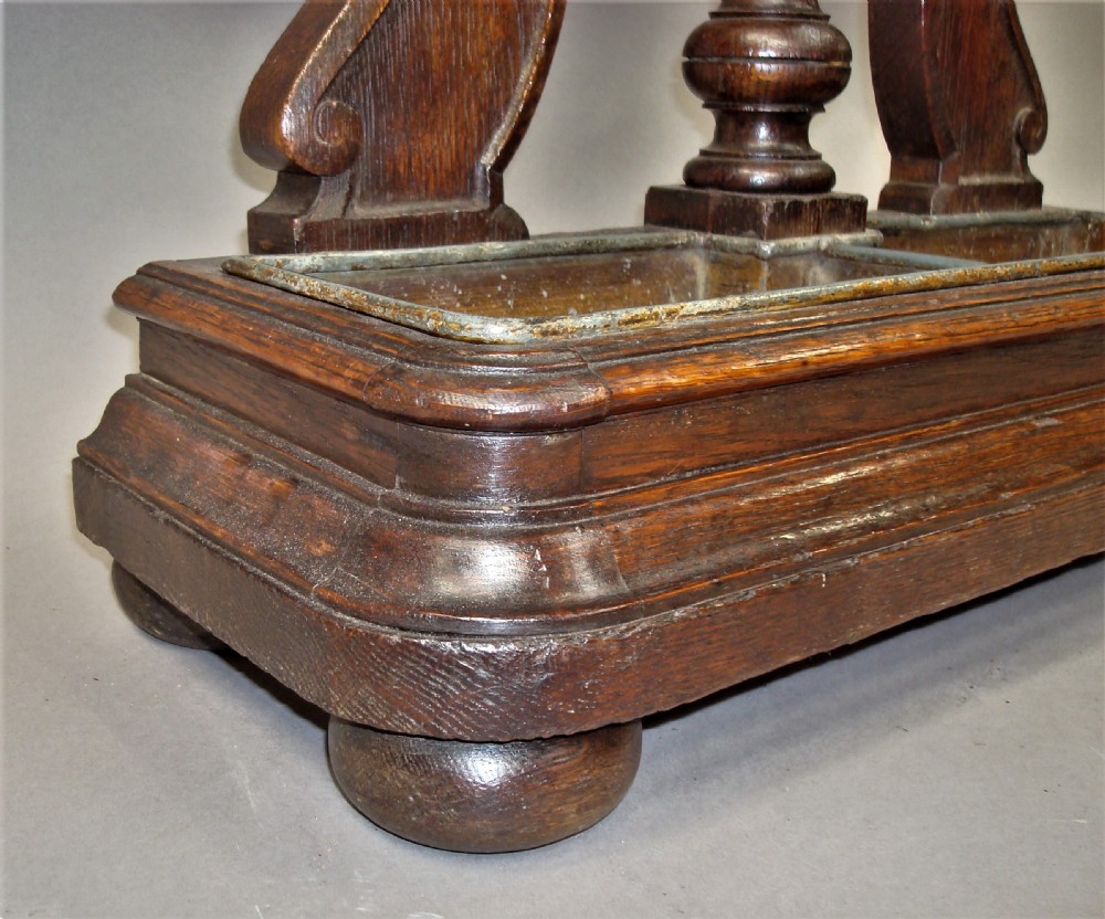 An antique dark oak stick stand featuring scroll support arms, a carved top cresting, a central column, and a removable metal drip tray at the base. View XIV