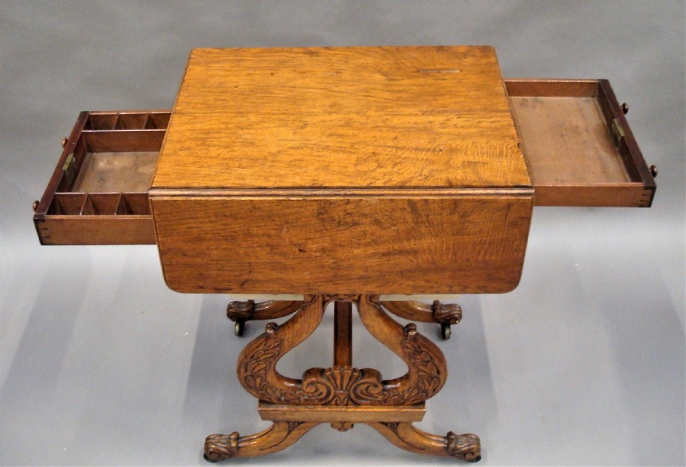 A Regency pollard oak work table featuring two drop flaps, two small drawers, a pleated fabric basket underneath, and carved scroll legs on brass castors. View IV