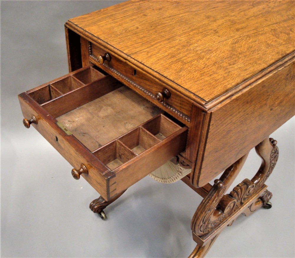 A Regency pollard oak work table featuring two drop flaps, two small drawers, a pleated fabric basket underneath, and carved scroll legs on brass castors. View V