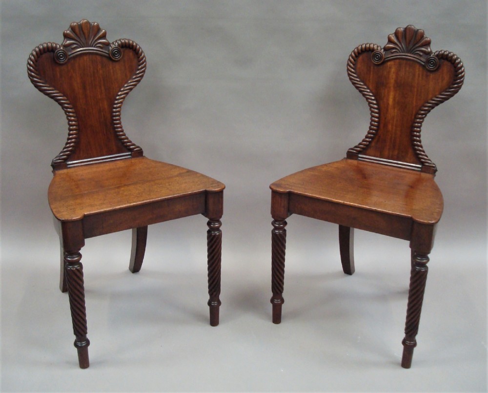 A pair of Regency mahogany hall chairs featuring carved shell cresting, rope-twist backrest borders, solid seats, and spiral-turned front legs.