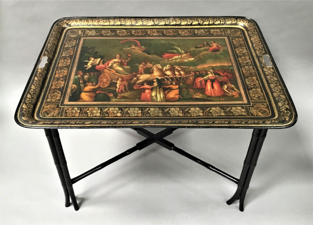 A large Regency painted toleware tray featuring a classical mythological scene and gilded borders, mounted on a black faux bamboo cross-leg stand.