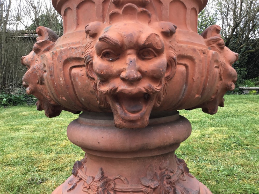 A nineteenth-century terracotta garden urn featuring sculpted mask faces around the body and floral swags on a pedestal base. View II