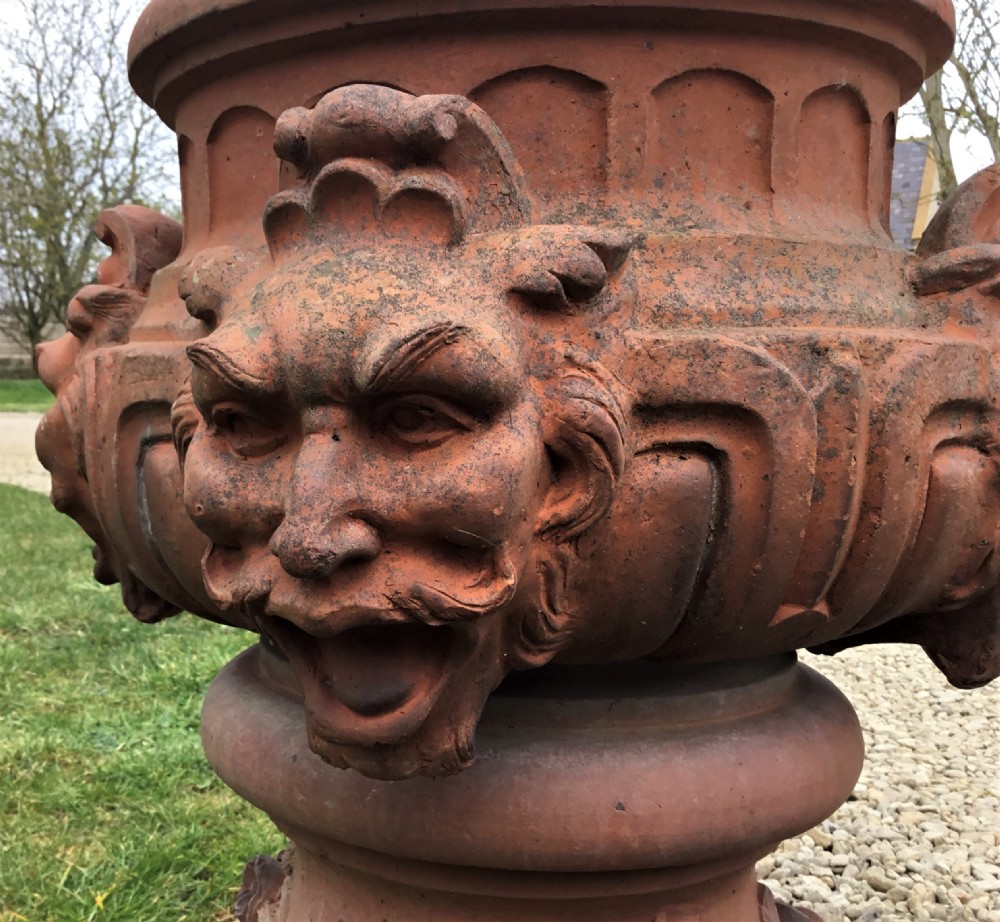 A nineteenth-century terracotta garden urn featuring sculpted mask faces around the body and floral swags on a pedestal base. View VII