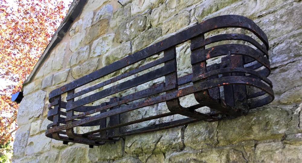A pair of early 20th-century black wrought iron window boxes with slatted horizontal straps, rounded ends, and top mounting hooks resting on gravel. View V