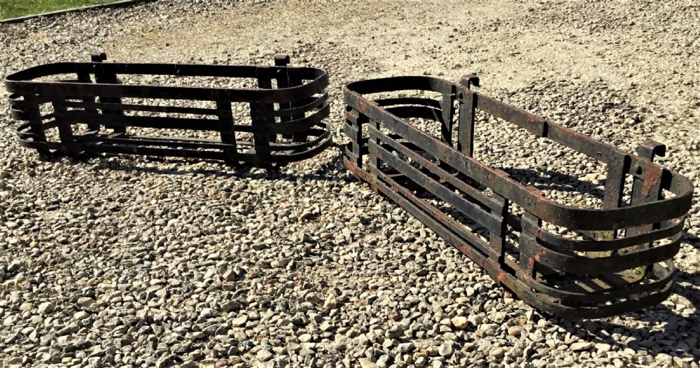 A pair of early 20th-century black wrought iron window boxes with slatted horizontal straps, rounded ends, and top mounting hooks resting on gravel.