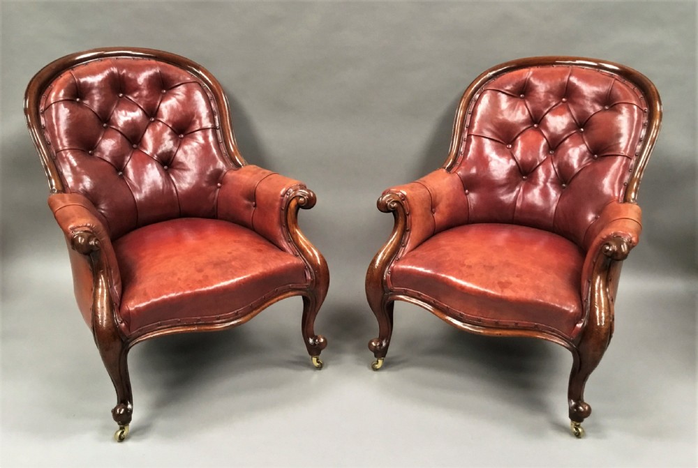 A pair of Victorian mahogany armchairs upholstered in button-backed deep red leather featuring scrolled arms and front cabriole legs on brass castors.