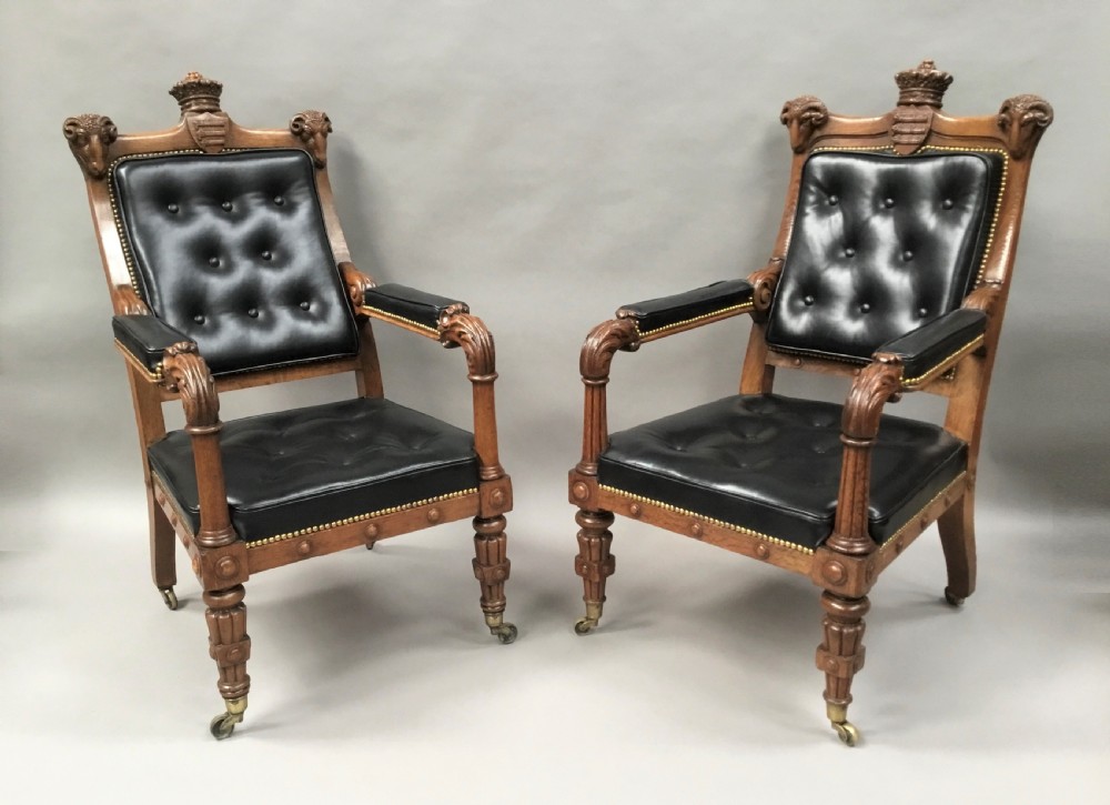 A pair of 19th-century carved oak library armchairs upholstered in buttoned black leather with detailed rams head finials and coronet cresting.