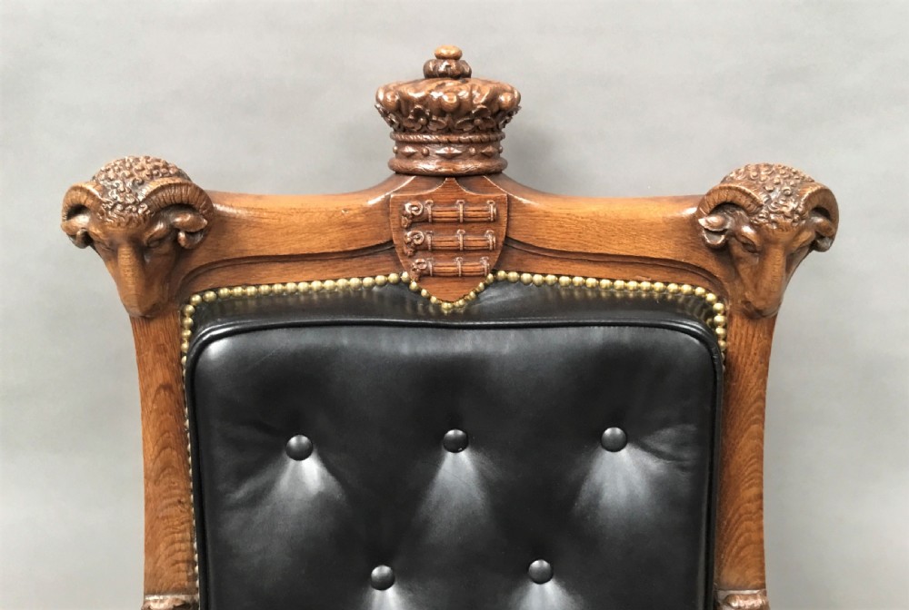 A pair of 19th-century carved oak library armchairs upholstered in buttoned black leather with detailed rams head finials and coronet cresting. View II