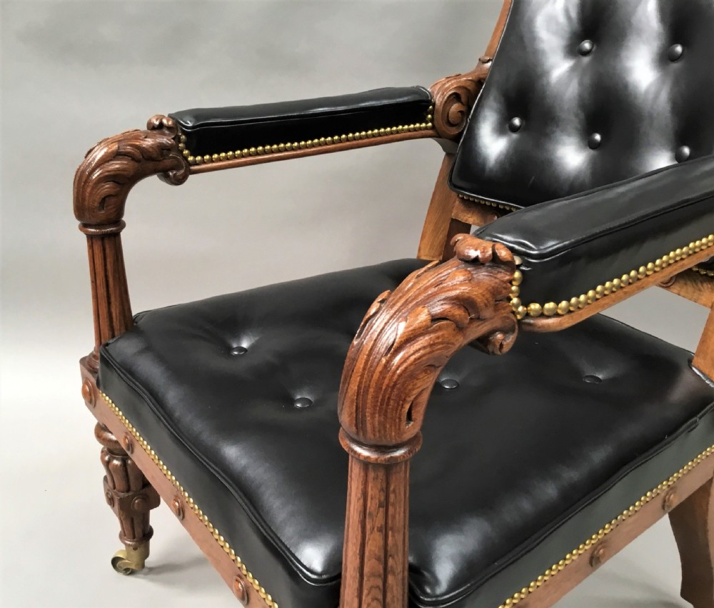 A pair of 19th-century carved oak library armchairs upholstered in buttoned black leather with detailed rams head finials and coronet cresting. View X