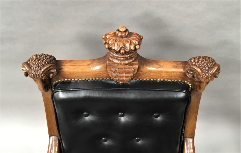 A pair of 19th-century carved oak library armchairs upholstered in buttoned black leather with detailed rams head finials and coronet cresting. View XVIII