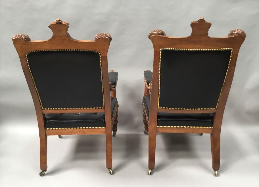 A pair of 19th-century carved oak library armchairs upholstered in buttoned black leather with detailed rams head finials and coronet cresting. View XIX