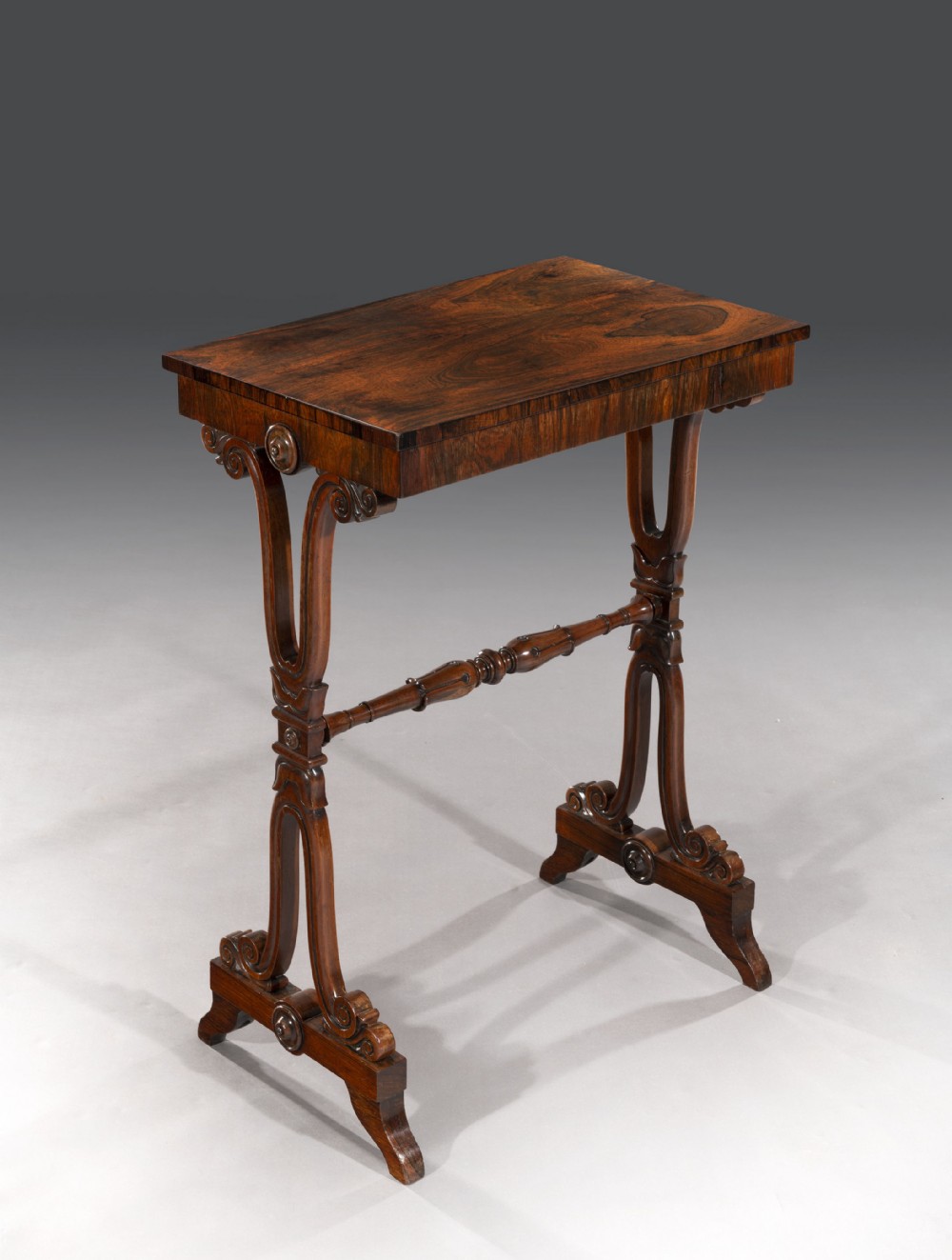 A Regency rosewood occasional table featuring a rectangular figured veneer top supported by scroll-shaped trestle legs with carved rosettes and a central turned stretcher.