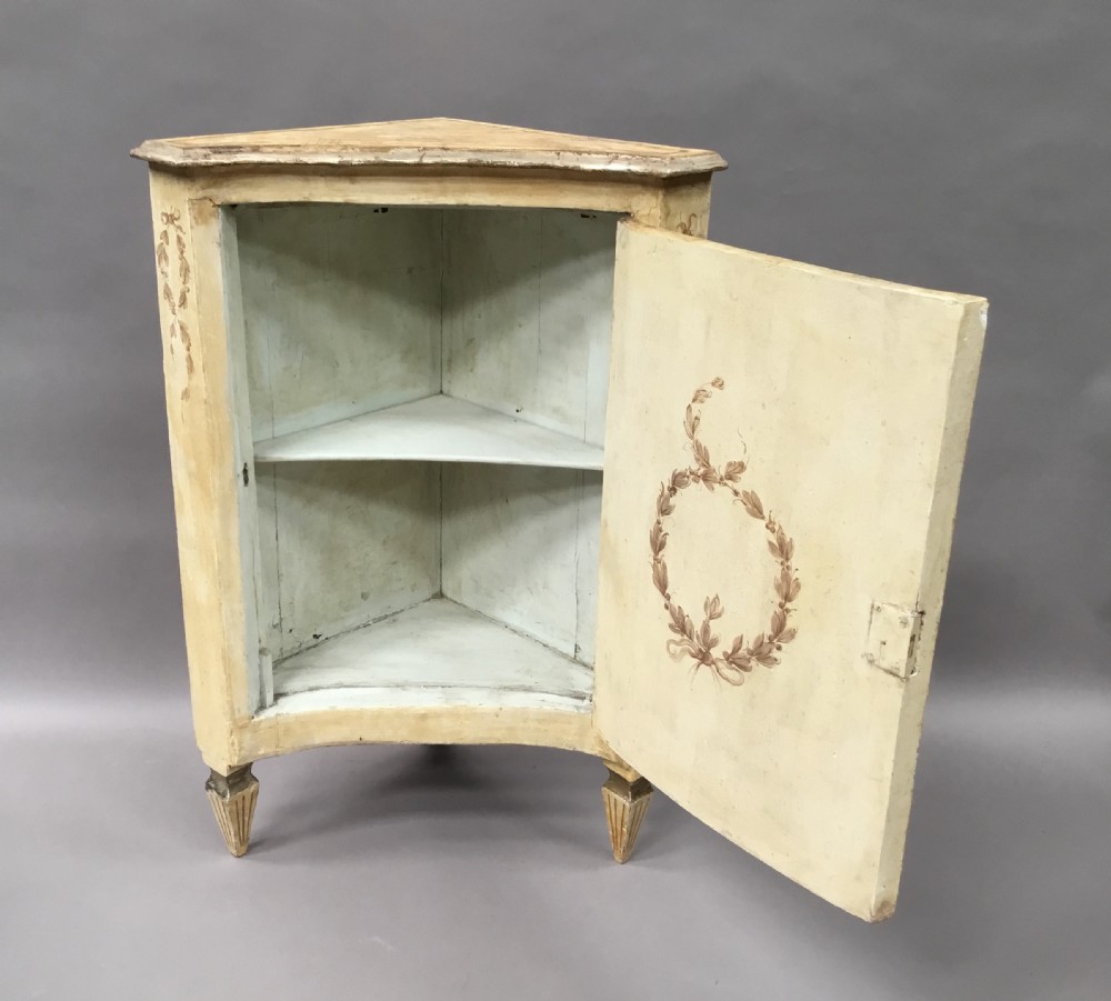 An 18th-century cream-painted Italian standing corner cabinet featuring a curved door decorated with a painted architectural scene and tapered legs. View X