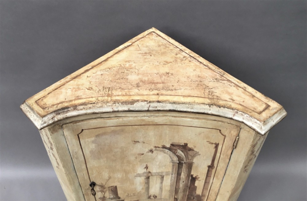An 18th-century cream-painted Italian standing corner cabinet featuring a curved door decorated with a painted architectural scene and tapered legs. View XIV