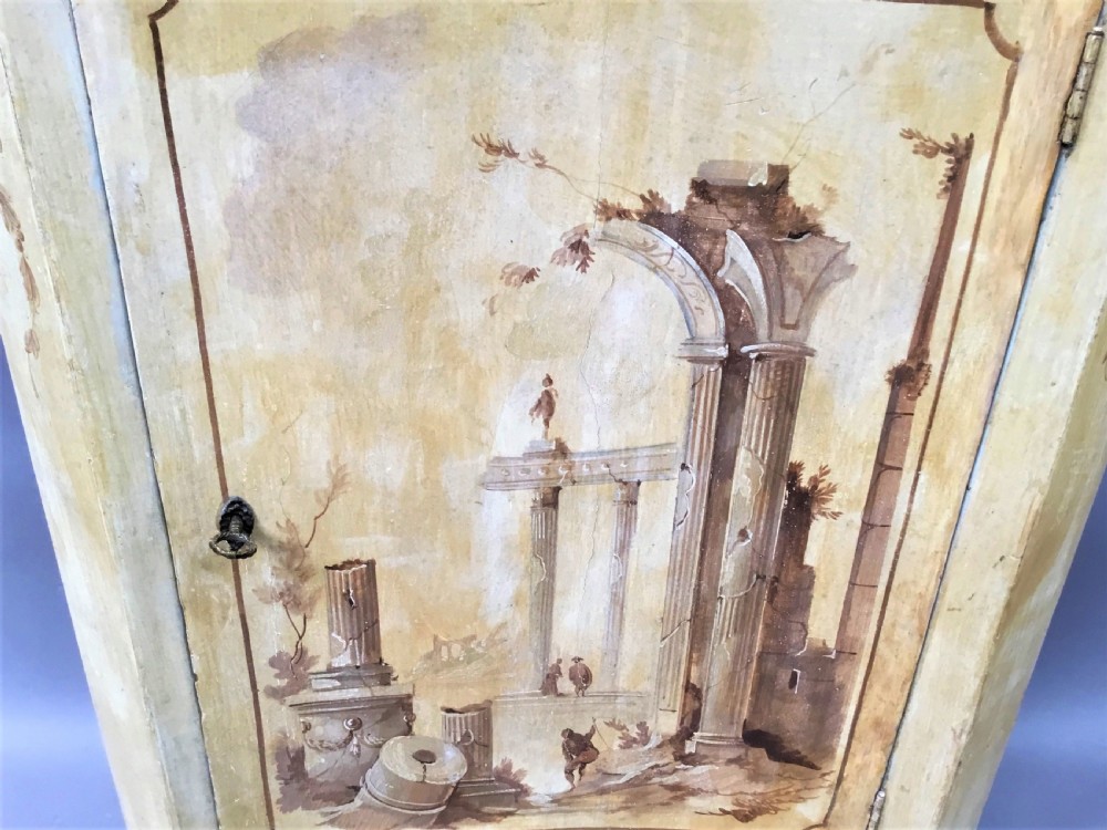 An 18th-century cream-painted Italian standing corner cabinet featuring a curved door decorated with a painted architectural scene and tapered legs. View XV