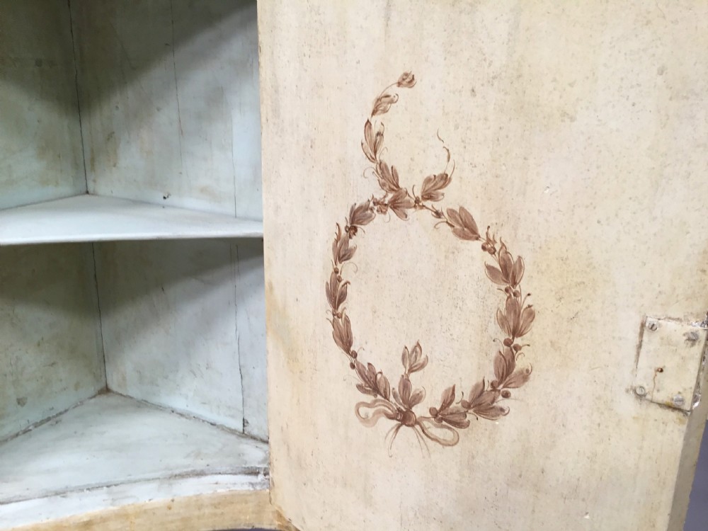 An 18th-century cream-painted Italian standing corner cabinet featuring a curved door decorated with a painted architectural scene and tapered legs. View XVI