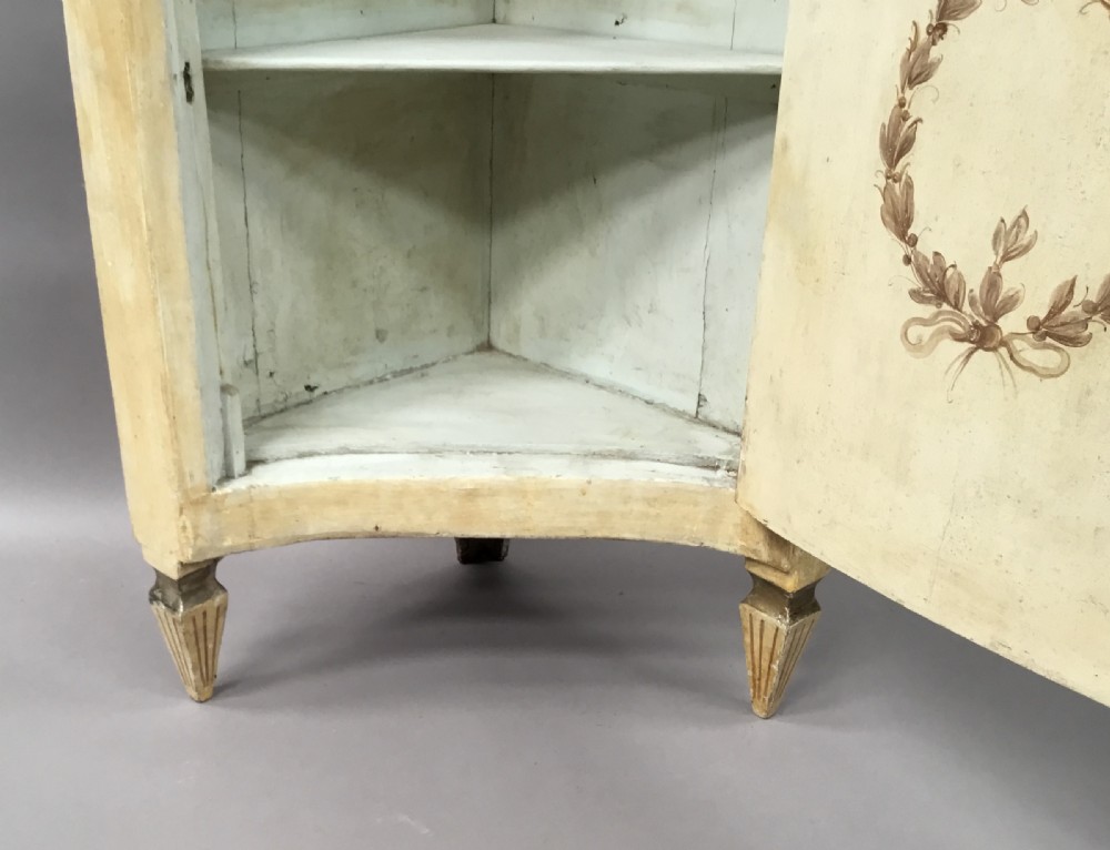 An 18th-century cream-painted Italian standing corner cabinet featuring a curved door decorated with a painted architectural scene and tapered legs. View XVII