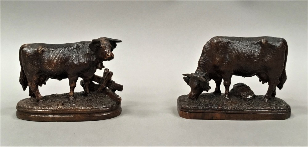 A pair of late nineteenth-century carved dark wood cows mounted on integrated plinths, showing textured coats and bells carved around their necks. View IX