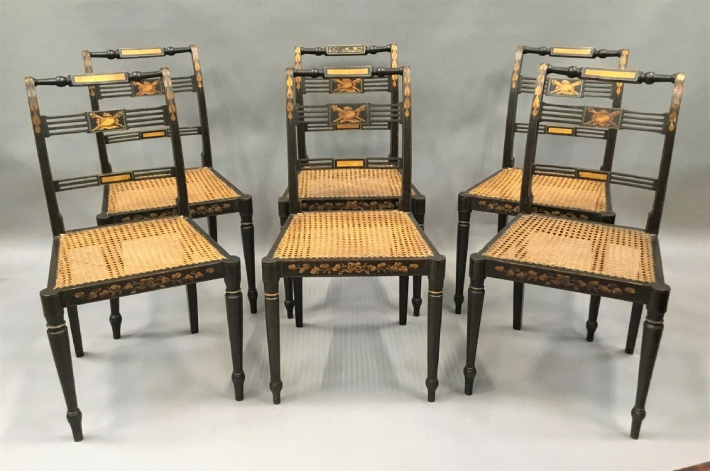 A set of six black-painted Regency side chairs featuring detailed parcel gilt decoration, horizontal slat backs, and natural woven cane seats.
