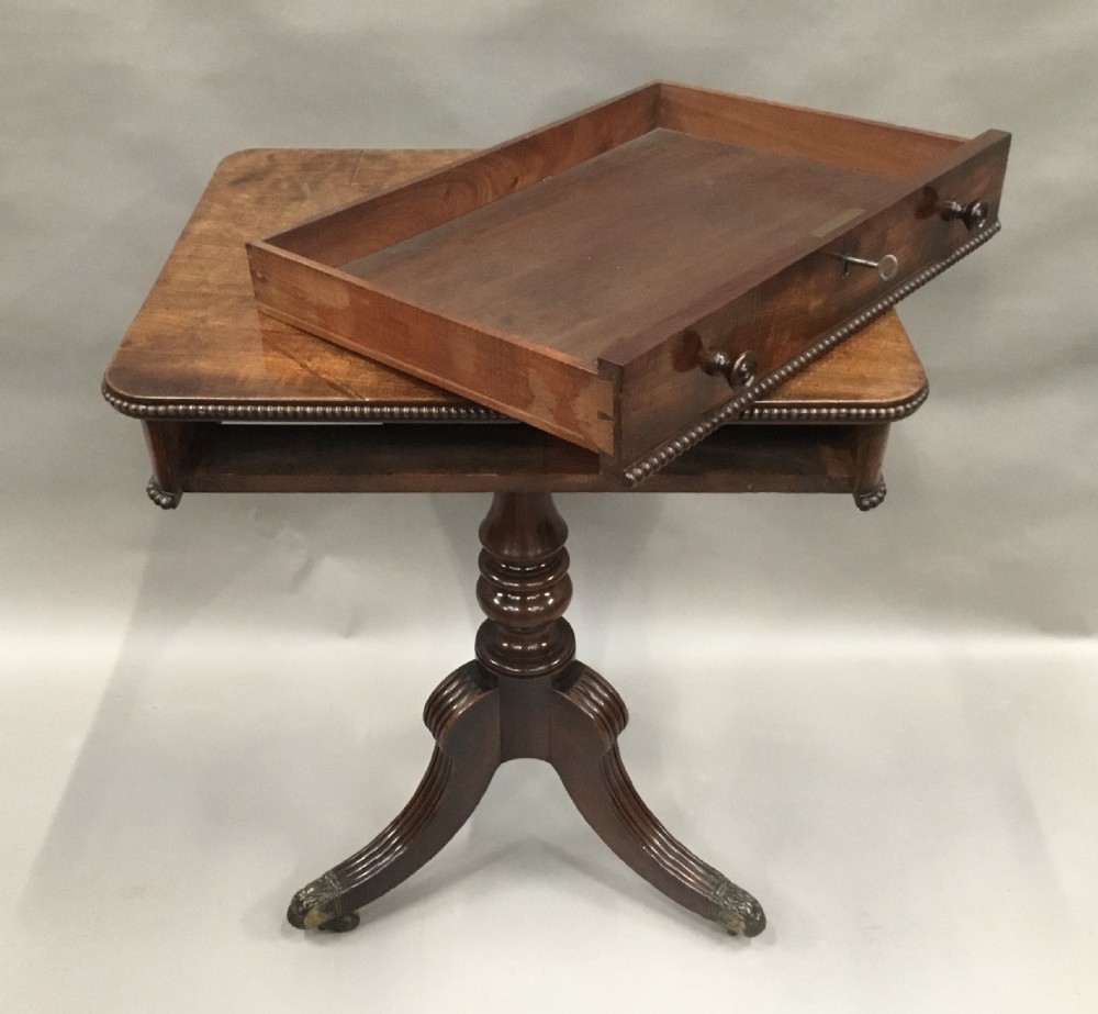 A Regency mahogany writing table featuring an adjustable hinged top, a single drawer with turned knobs, a turned central pedestal, and reeded tripod legs ending in brass castors. View XVIII