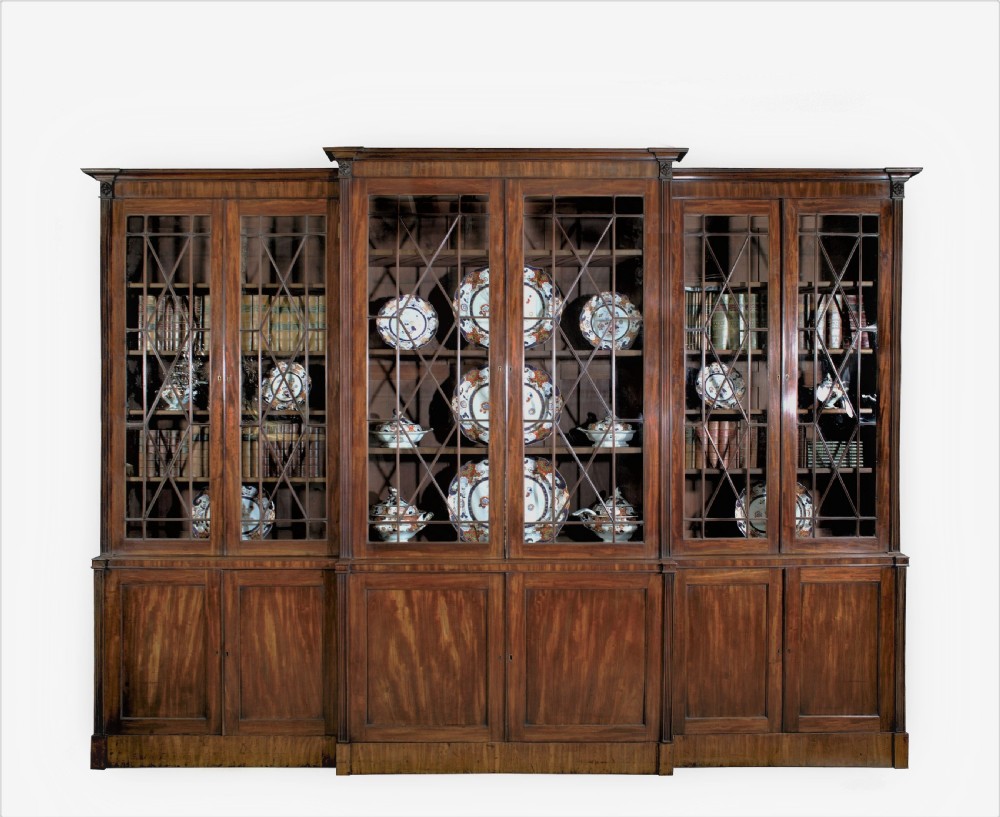 A large George III mahogany breakfront bookcase with a central pediment, glazed upper doors with geometric glazing bars, and flame mahogany cupboard doors below. View II
